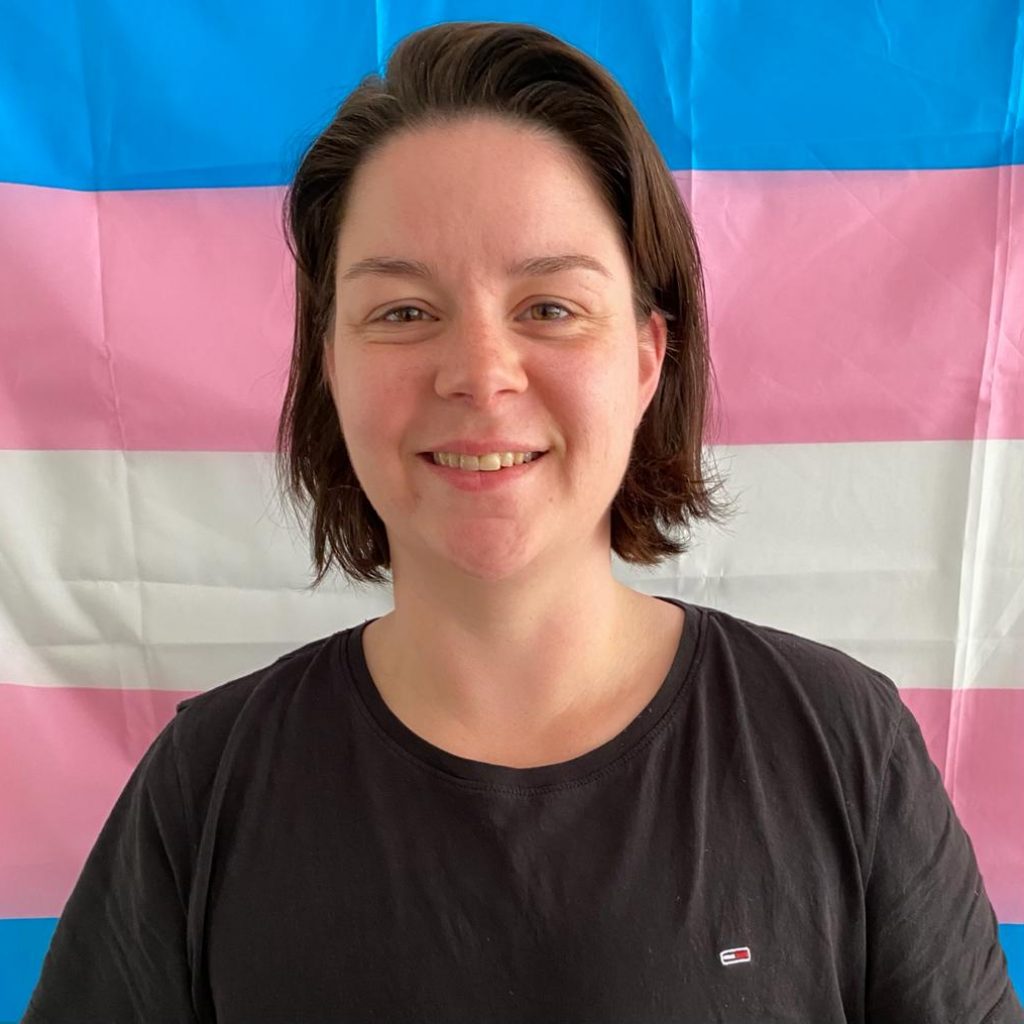 Een foto van Ing. Die staat voor een transgender vlag. Ing heeft bruin haar, lichtbruine ogen en heeft een zwart shirt aan.