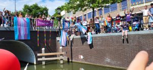 Deelnemers en vrijwilligers van Transcafé LVB zwaaien vanaf de kant naar een boot tijdens de Utrecht Canal pride 2022.