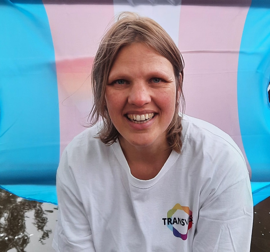 Een foto van Anne. Anne staat wat gebogen voor een transgendervlag en heeft een wit t-shirt met Transvisie logo aan. Anne heeft blond haar en een brede glimlach.